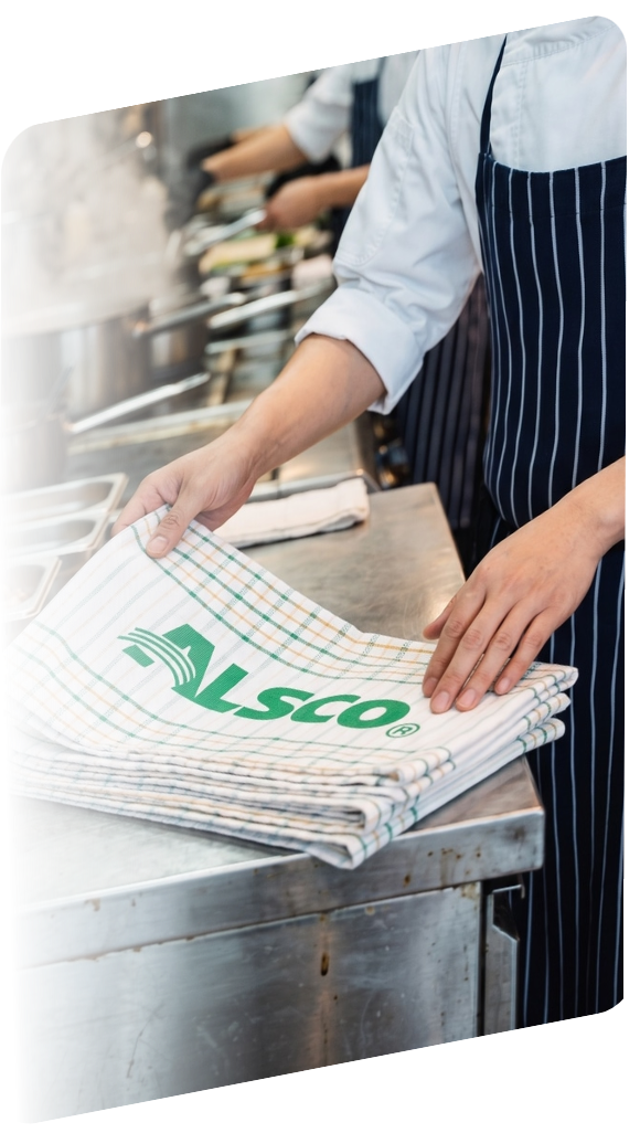 Chef in a striped apron folding a stack of Alsco tea towels on a stainless steel kitchen bench