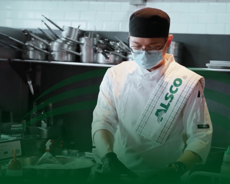 Chef in a white chef jacket and face mask with an Alsco tea towel over his shoulder in a commercial kitchen