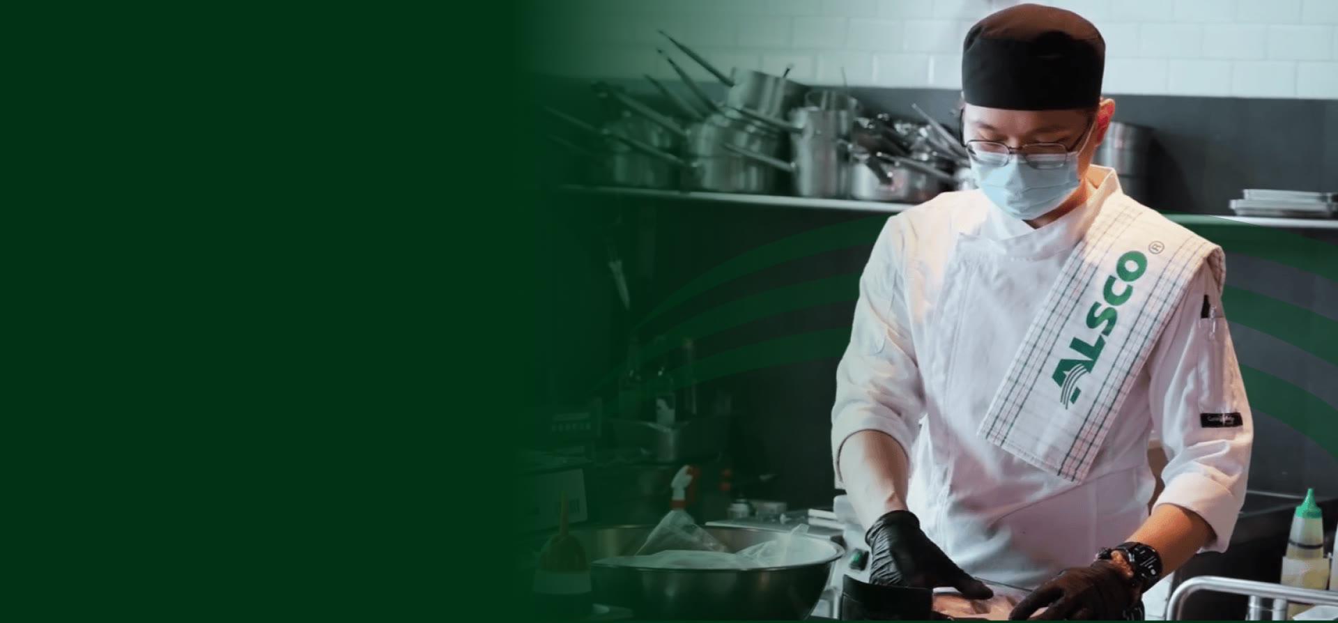 Chef in a white chef jacket and face mask with an Alsco tea towel over his shoulder in a commercial kitchen