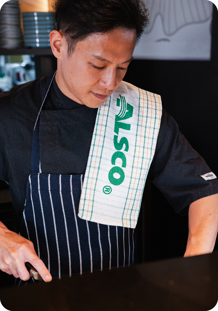 Chef in a black chef jacket and striped apron with an Alsco tea towel over his shoulder