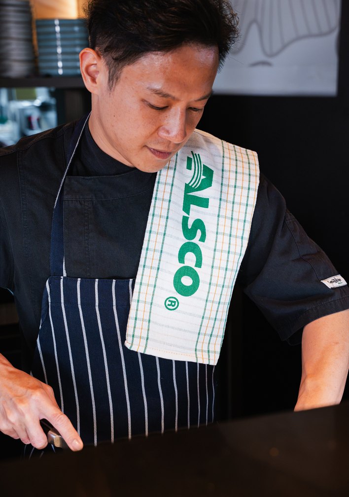 Chef in a black chef jacket and striped apron with an Alsco tea towel over his shoulder