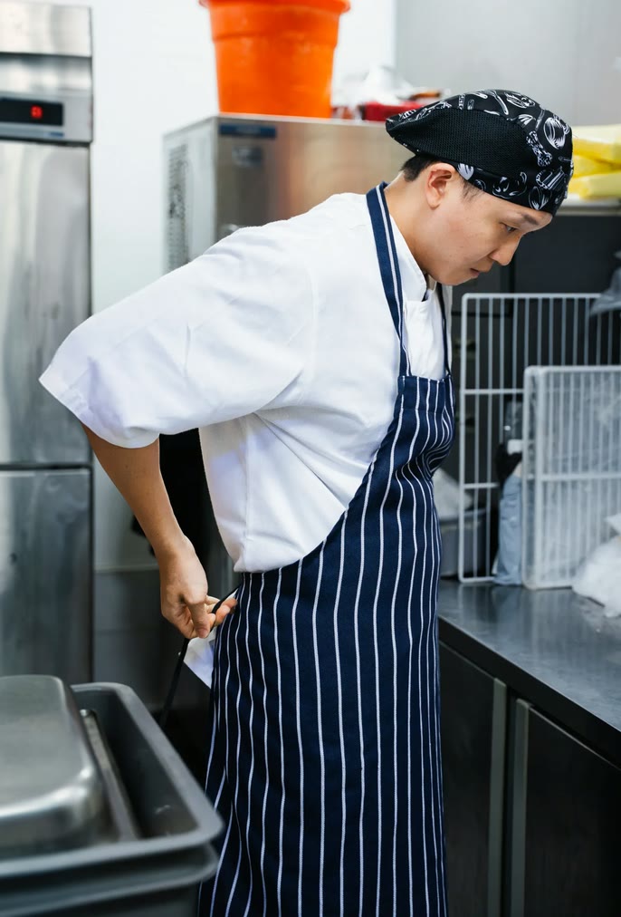 Chef in a white jacket and navy striped apron working in a stainless steel commercial kitchen