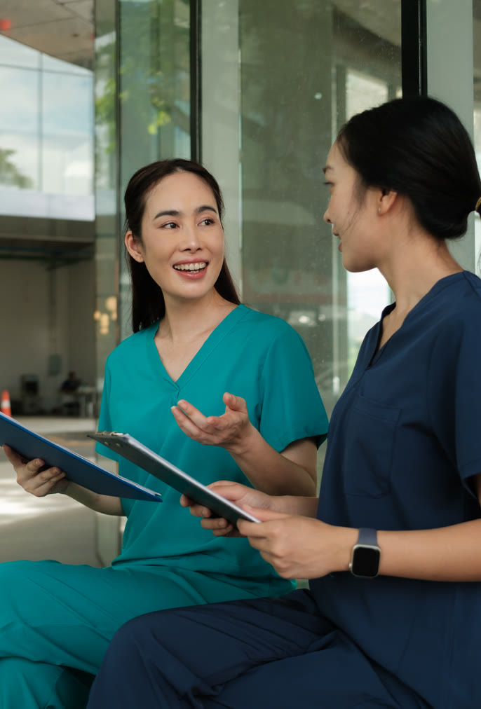 Two healthcare workers in teal and navy scrubs discussing notes on clipboards