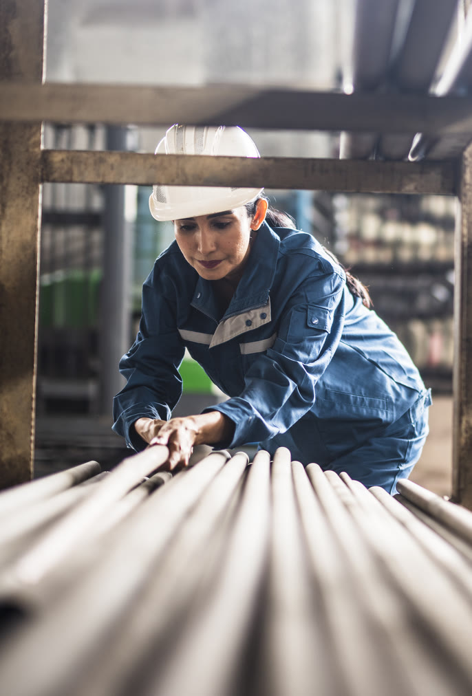 Worker in a white hard hat and navy coveralls handling steel rods in a manufacturing plant